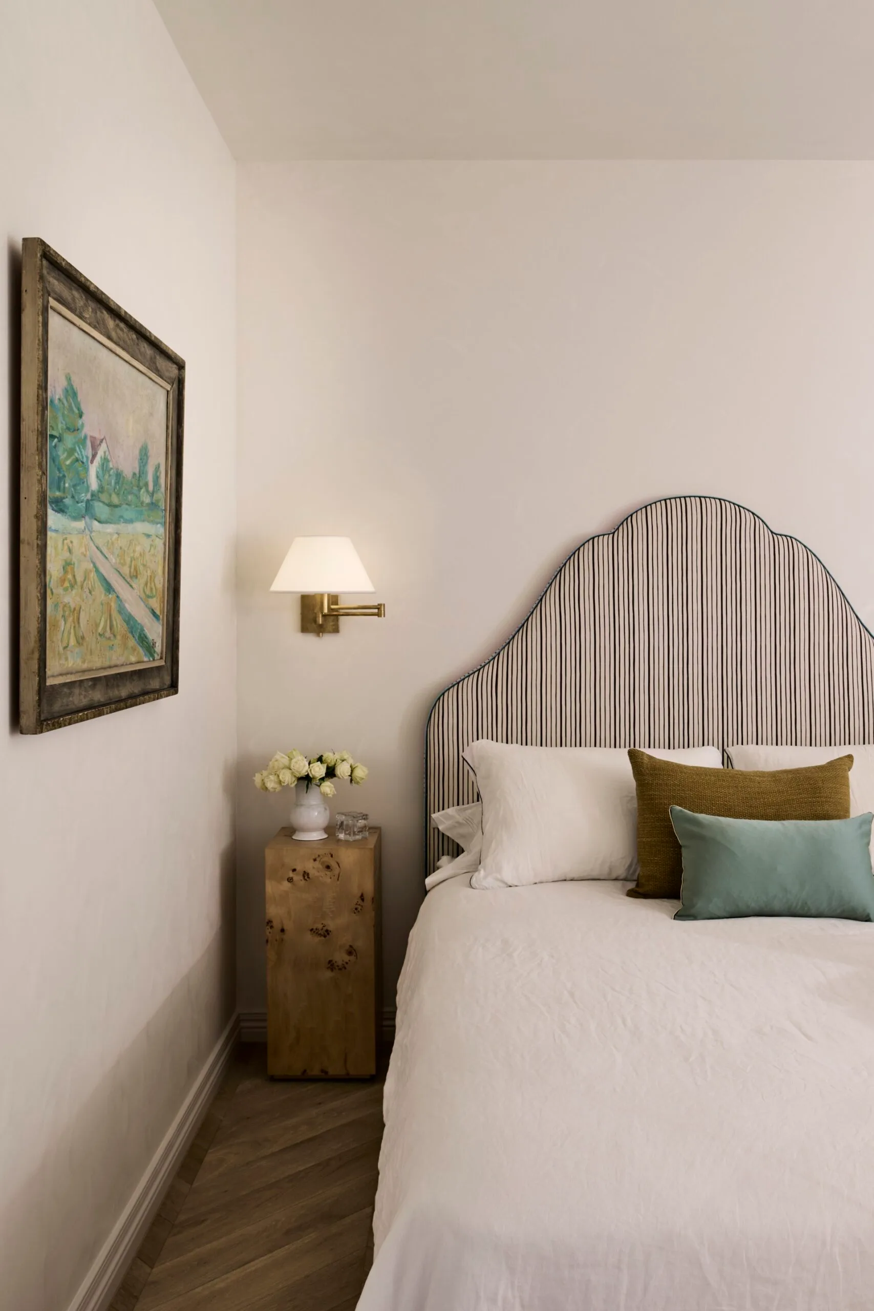 Cosy bedroom with a striped curved headboard, white bedding, decorative pillows, bedside table with flowers underneath a wall-mounted sconce with white shade, and an antique painting mounted on the wall to the left of the bed.