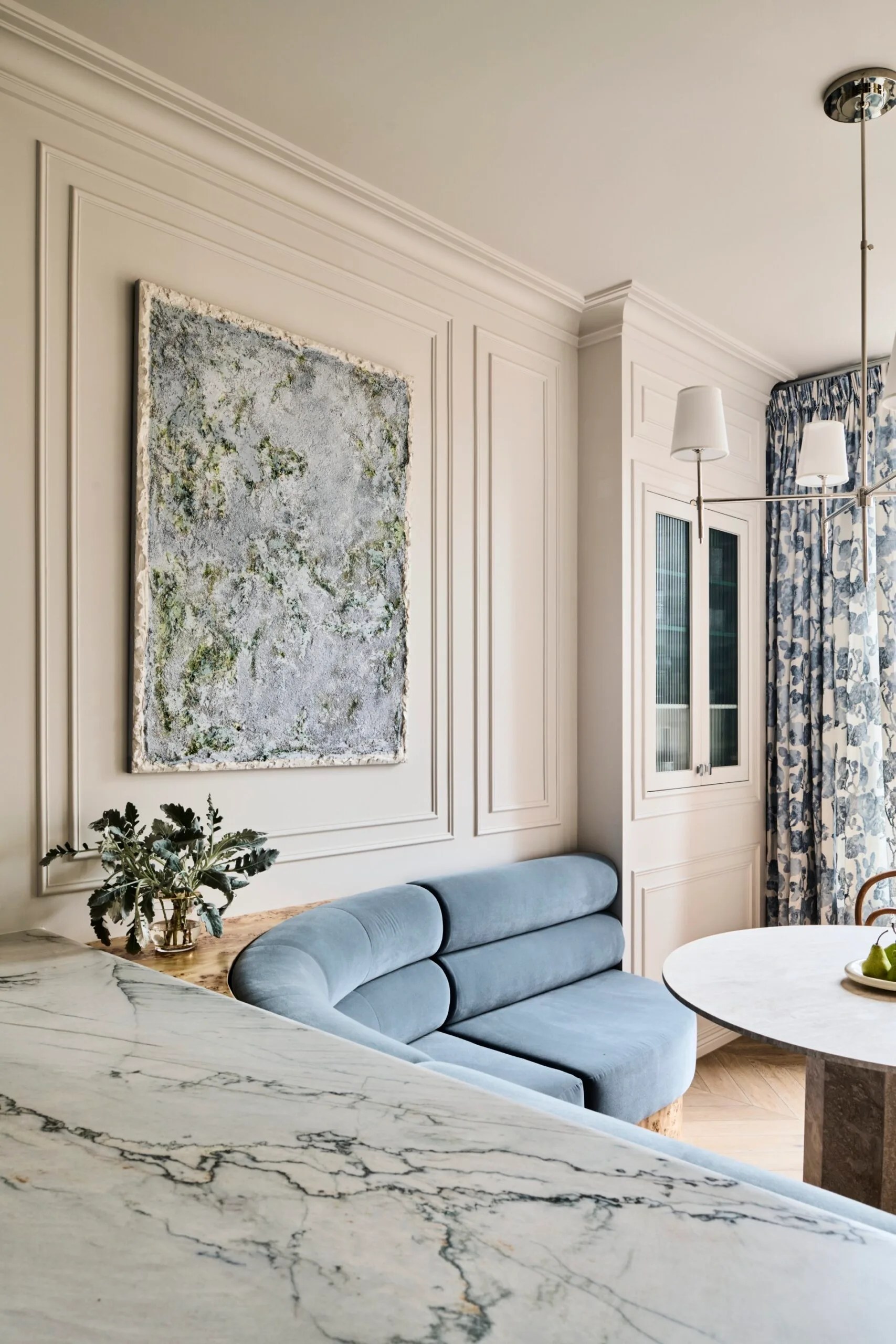 Stylish dining area in an apartment with blue built-in banquette, abstract painting positioned in the middle of decorative panelling on the white walls, a marble table, and floral drapes.