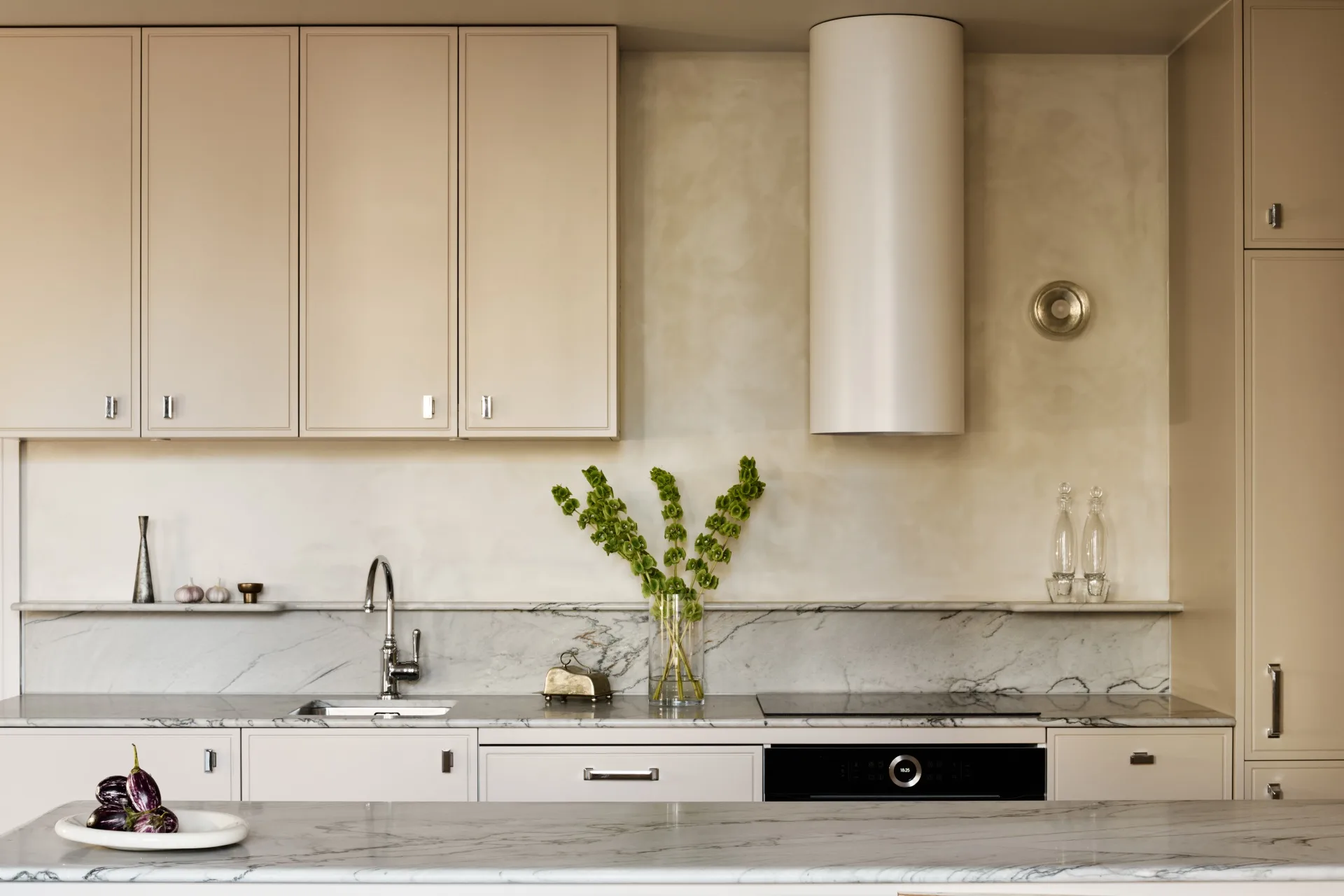 An apartment kitchen in a small apartment with beige cabinets, microcement walls, marble countertops and splashback, a vase of green flowers, and a bowl of eggplants. The top of a small island is in the foreground, with a countertop along the wall behind. A curved rangehood cover is also on the wall.