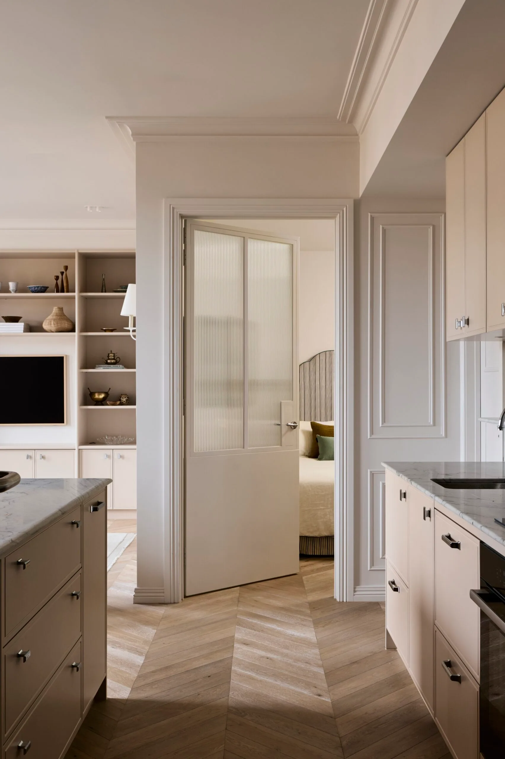 Neutral-toned kitchen in an apartment with marble countertops, open shelving, timber flooring laid in a chevron pattern and a view into a bedroom through a partially open door. To the left of the photo, open shelves and a tv are visible on a wall in the background.