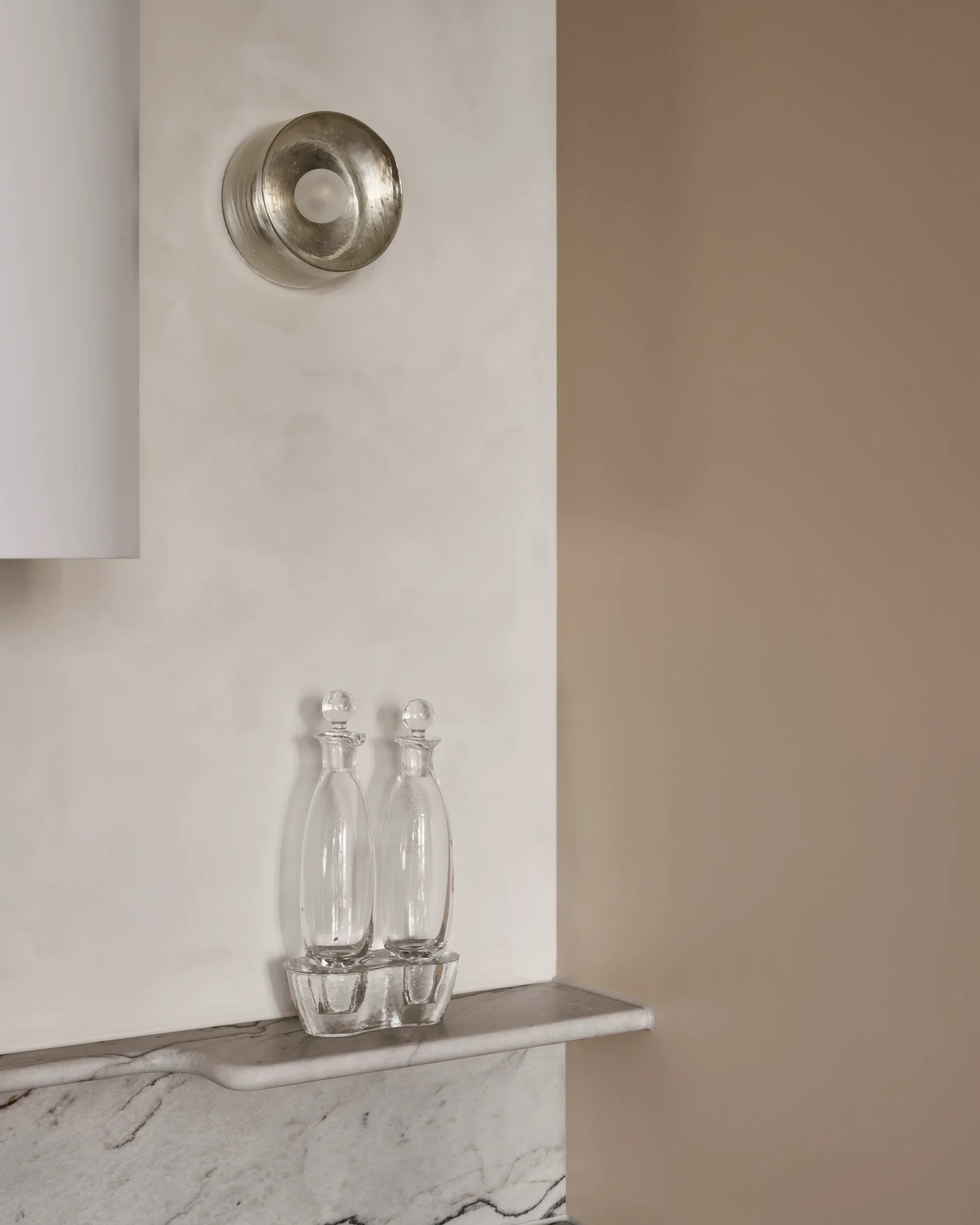 A close up of beige microcement walls, with a round wall sconce attached. At the bottom of the photo, a marble splashback leads up to a small marble ledge. Two vintage oil bottles are displayed on the ledge.