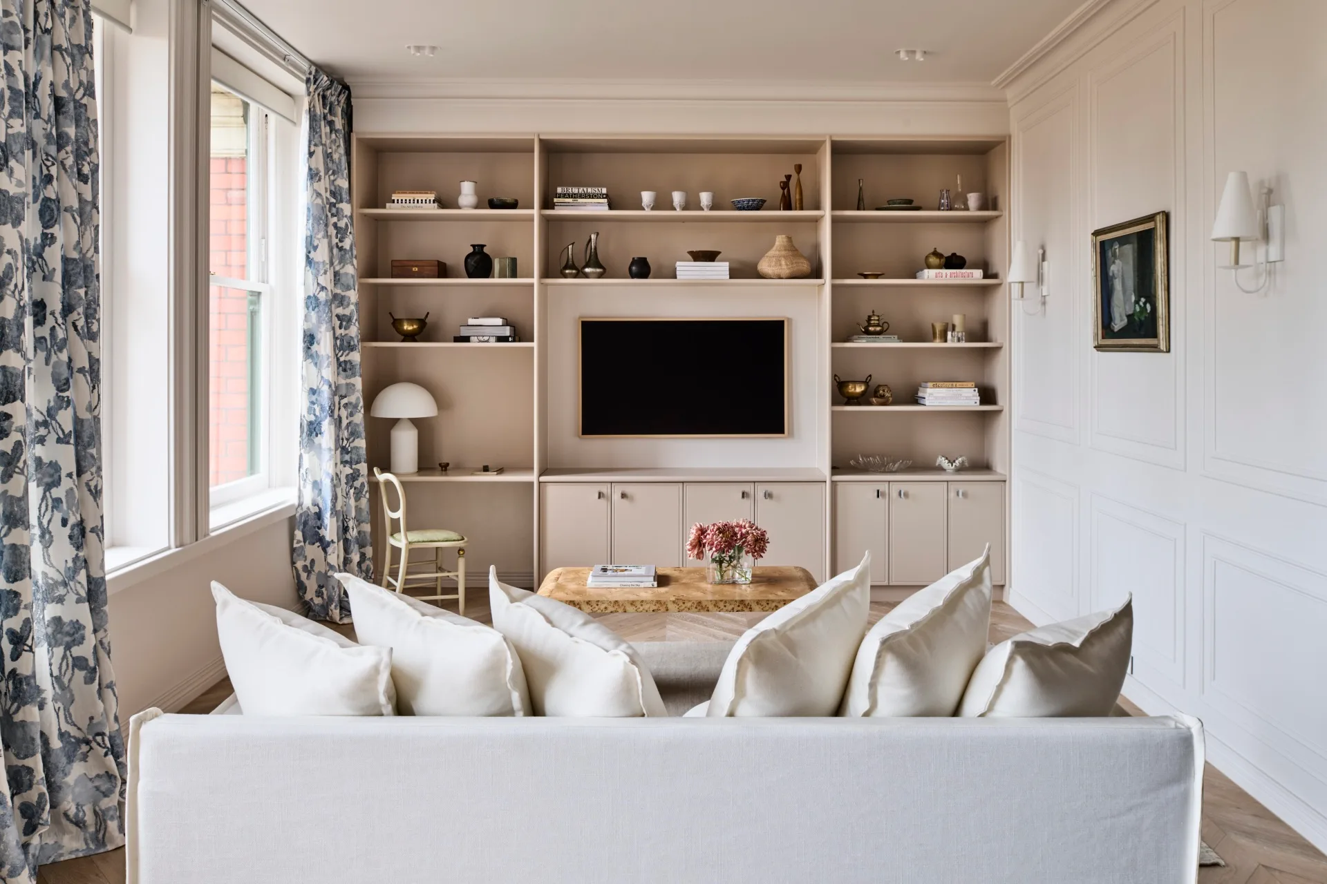 Living room in an apartment. The back of a white sofa with plentiful cushions atop is visible from the back in the foreground. It faces a wall of built-in joinery which has a television mounted on the wall in the middle. Floral curtains are positioned to the left, while along the right wall is decorative panelling, an antique paining and two wall sconces with white lampshades.