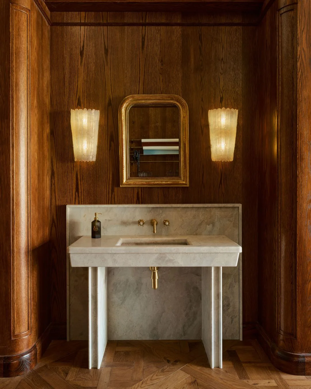 A deep brown timber wall with a freestanding marble vanity and basin, with gold tapware, a mirror above, and two antique wall sconces for light at the Libertine Parfumerie store in Melbourne