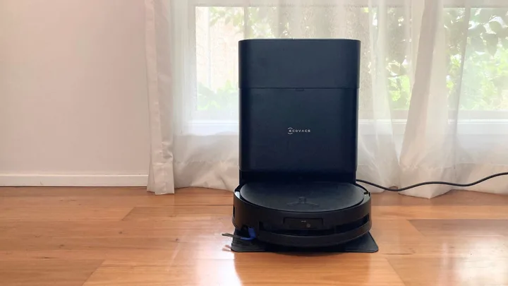 An Ecovacs robot vacuum cleaner and mop in the docking station. Both are black. They are on wooden floorboards, in front of a window with a white sheer curtain.