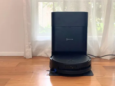 An Ecovacs robot vacuum cleaner and mop in the docking station. Both are black. They are on wooden floorboards, in front of a window with a white sheer curtain.