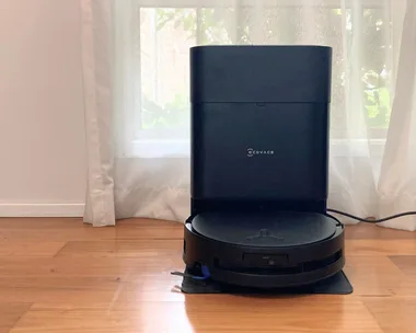 An Ecovacs robot vacuum cleaner and mop in the docking station. Both are black. They are on wooden floorboards, in front of a window with a white sheer curtain.