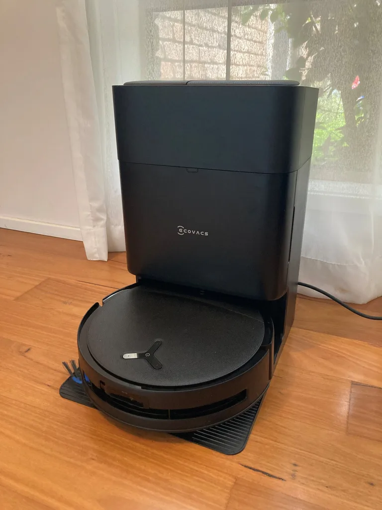 An Ecovacs robot vacuum cleaner and mop in the docking station. Both are black. They are on wooden floorboards, in front of a window with a white sheer curtain.