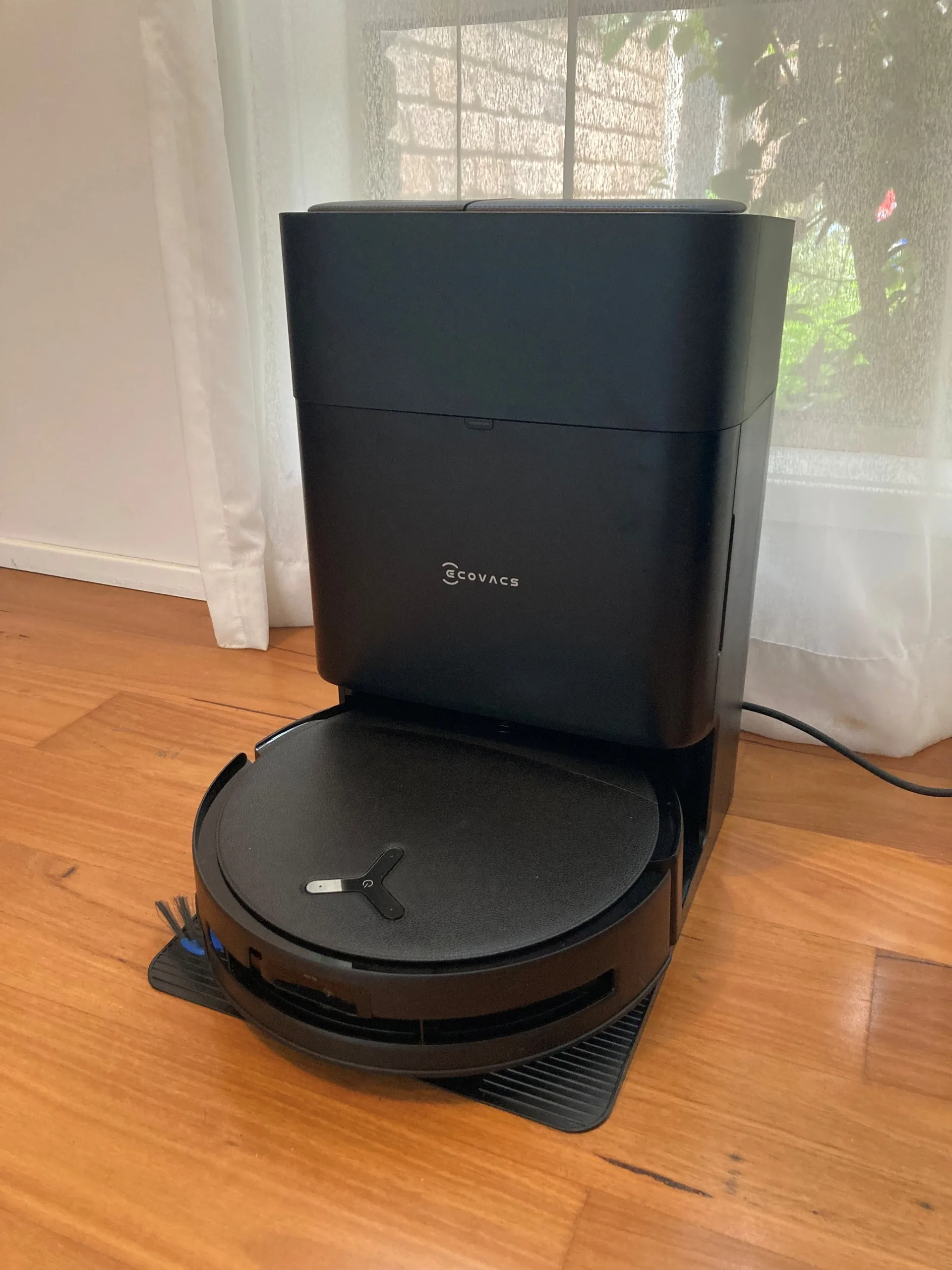 An Ecovacs robot vacuum cleaner and mop in the docking station. Both are black. They are on wooden floorboards, in front of a window with a white sheer curtain.