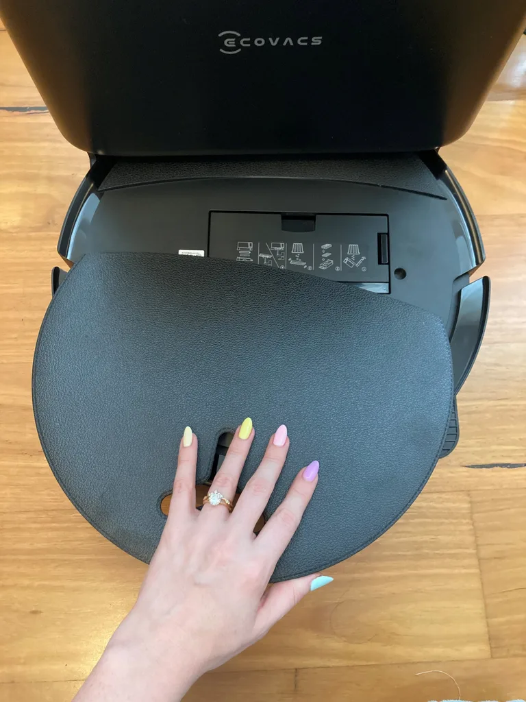 Photo of a robot vacuum cleaner. A hand is removing the lid, revealing instructions underneath.