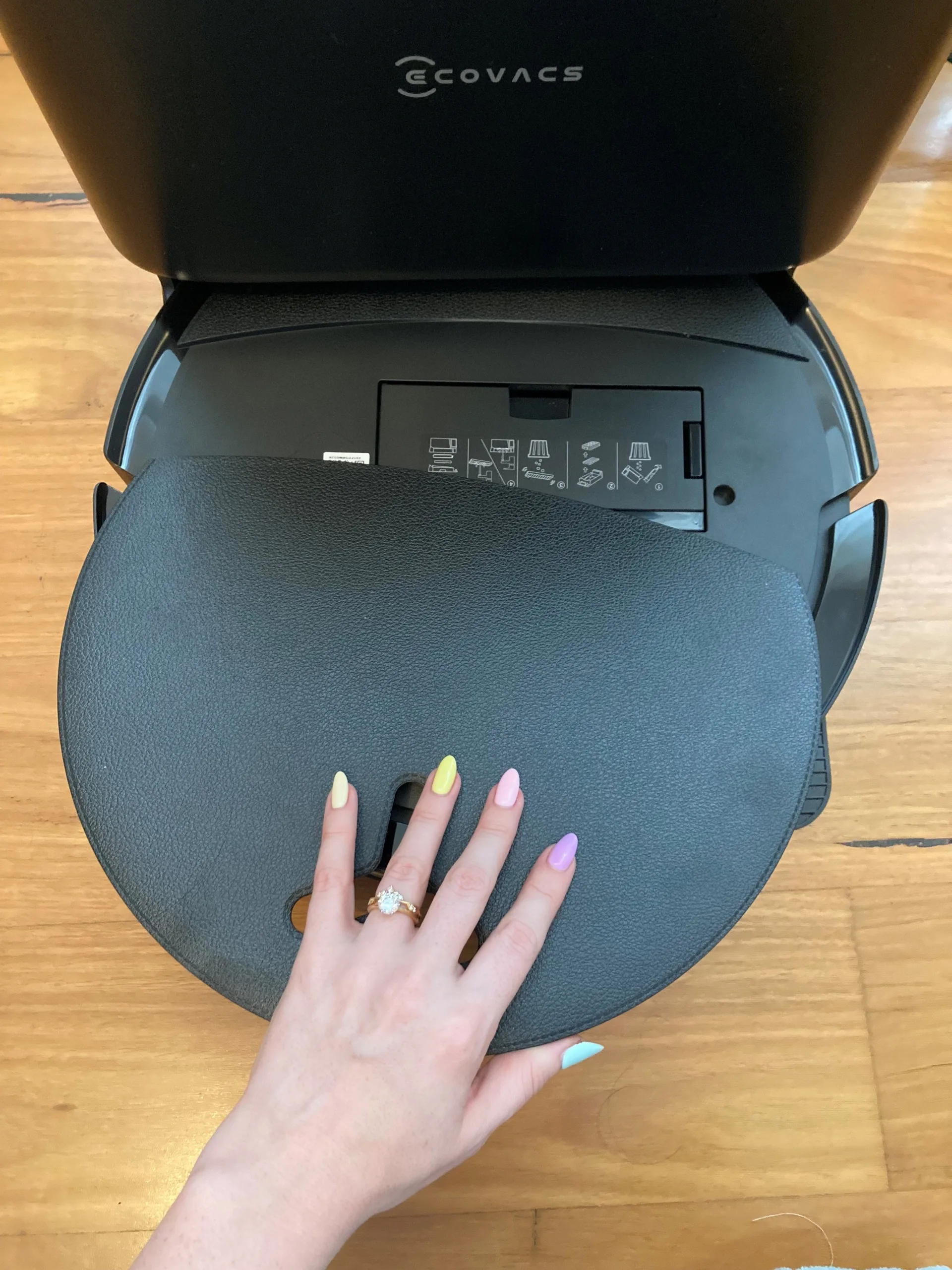 Photo of a robot vacuum cleaner. A hand is removing the lid, revealing instructions underneath.