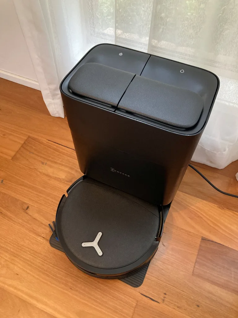 An Ecovacs robot vacuum cleaner and mop in the docking station. Both are black. They are on wooden floorboards, in front of a window with a white sheer curtain.