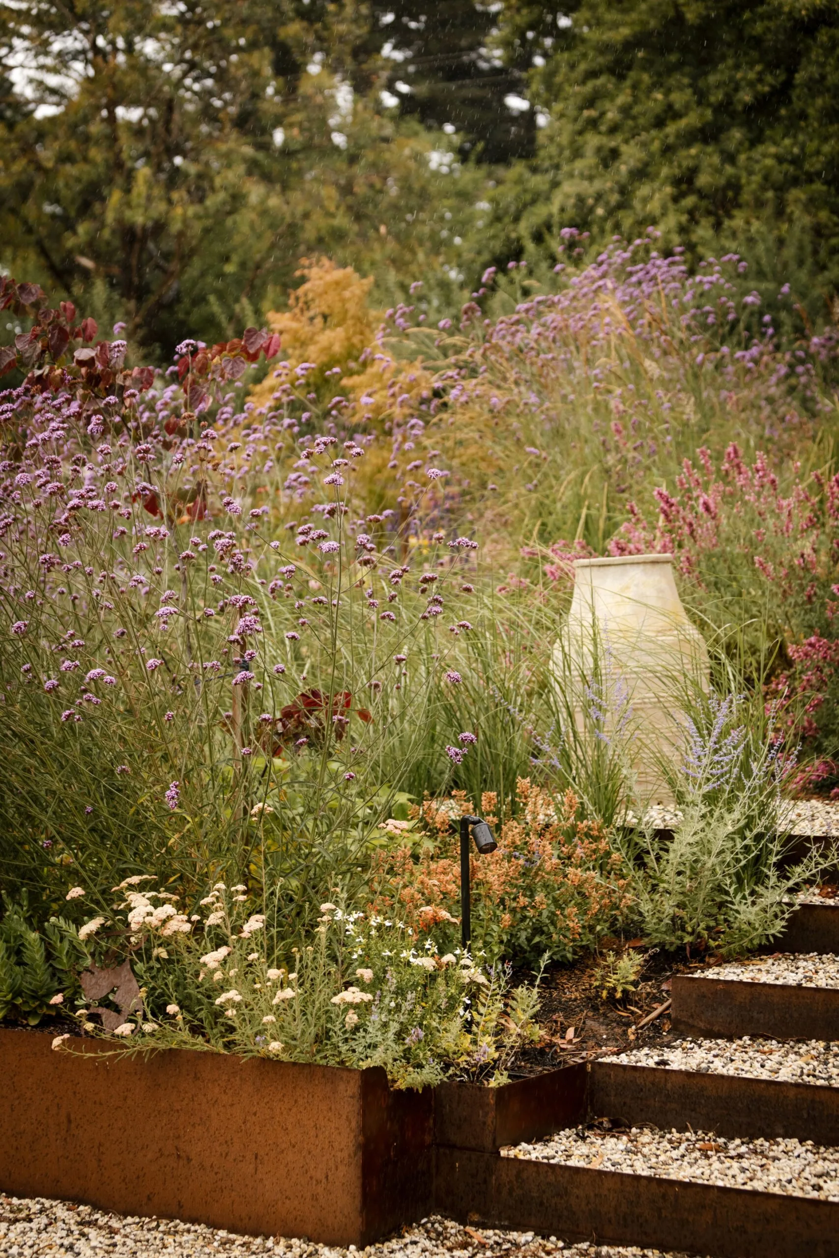 Garden with purple and white flowers, a stone pathway, and a large ceramic vase amidst lush greenery.