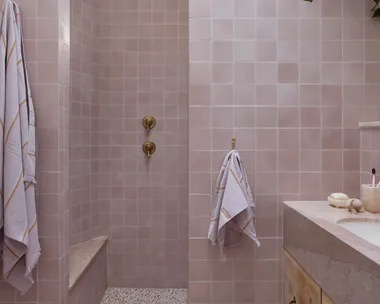 Pink tiled bathroom with terrazzo floor, wooden vanity, gold fixtures, and hanging plant. Two towels are on hooks.