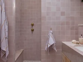 Pink tiled bathroom with terrazzo floor, wooden vanity, gold fixtures, and hanging plant. Two towels are on hooks.