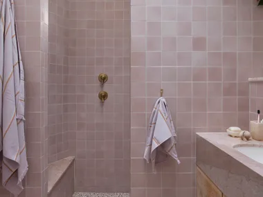 Pink tiled bathroom with terrazzo floor, wooden vanity, gold fixtures, and hanging plant. Two towels are on hooks.