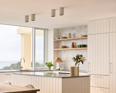 Modern kitchen with white cabinets, wooden dining table, and large windows offering a view of greenery outside.