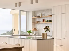 Modern kitchen with white cabinets, wooden dining table, and large windows offering a view of greenery outside.