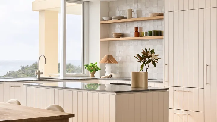 Modern kitchen with white cabinets and open shelves with colourful glass cups, wooden dining table, and large windows offering a view of greenery outside.