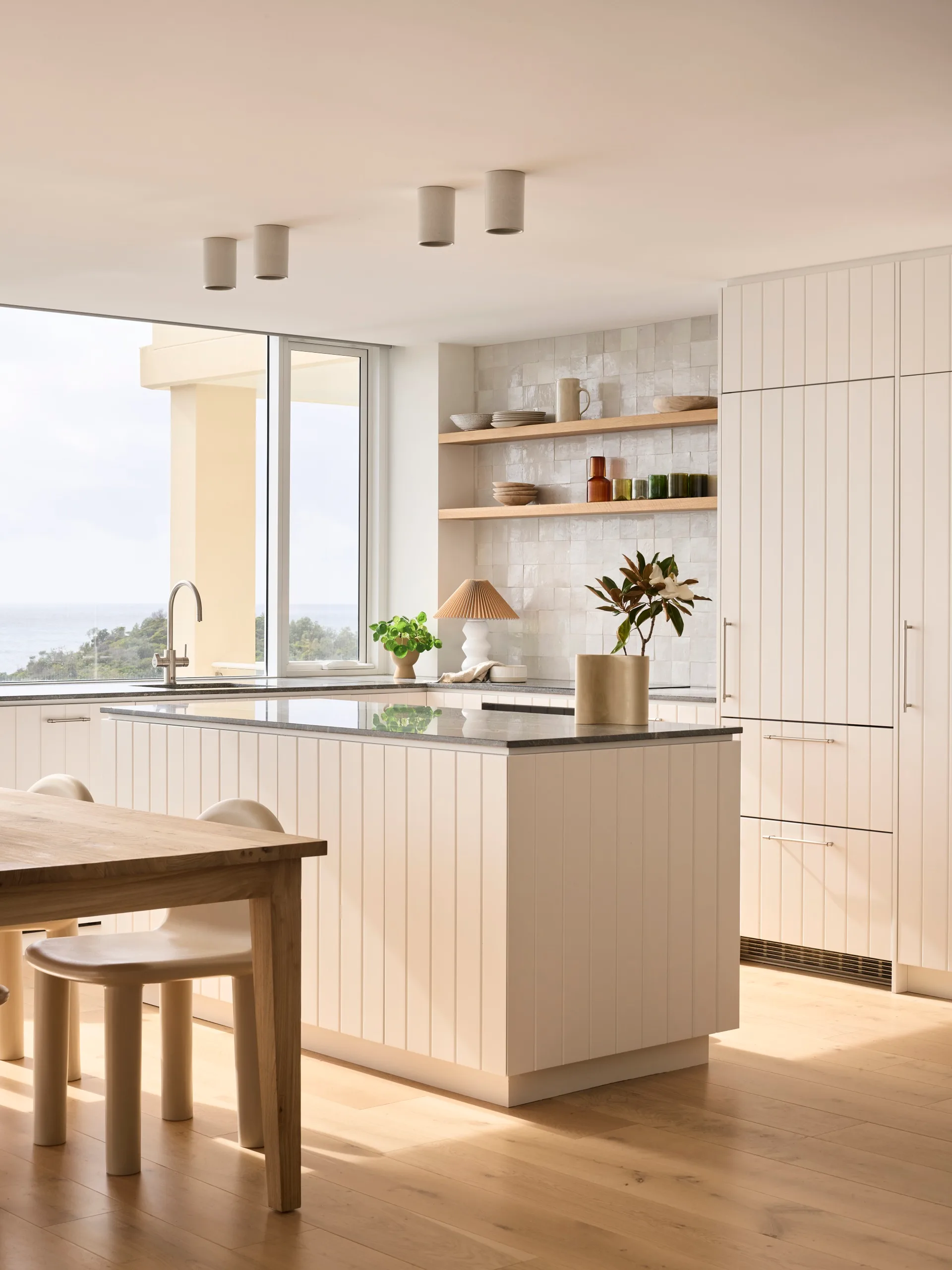 Modern kitchen with white cabinets, wooden dining table, and large windows offering a view of greenery outside.