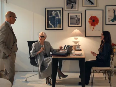 A still from The Devil Wears Prada 2, featuring Anne Hathaway as Andy Sachs, Stanley Tucci as Nigel Kipling and Meryl Streep as Miranda Priestly. They are in Miranda's office, which features white walls, framed photographs on the walls and a light timber and black desk with a white lamp on top. A chrome bar cart is to the side.