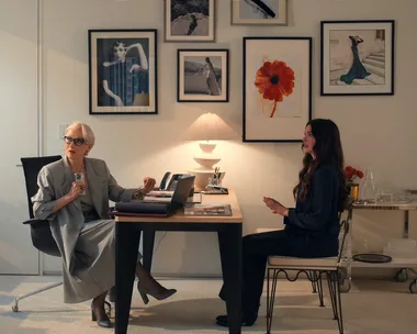 A still from The Devil Wears Prada 2, featuring Anne Hathaway as Andy Sachs, Stanley Tucci as Nigel Kipling and Meryl Streep as Miranda Priestly. They are in Miranda's office, which features white walls, framed photographs on the walls and a light timber and black desk with a white lamp on top. A chrome bar cart is to the side.