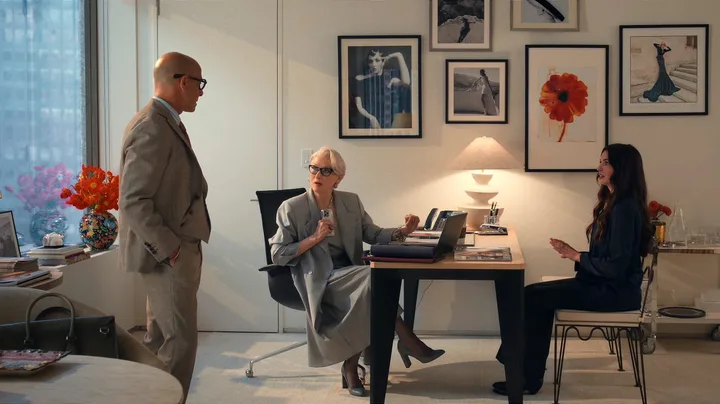 A still from The Devil Wears Prada 2, featuring Anne Hathaway as Andy Sachs, Stanley Tucci as Nigel Kipling and Meryl Streep as Miranda Priestly. They are in Miranda's office, which features white walls, framed photographs on the walls and a light timber and black desk with a white lamp on top. A chrome bar cart is to the side.