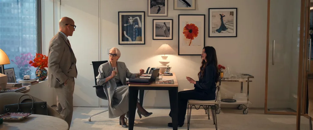 A still from The Devil Wears Prada 2, featuring Anne Hathaway as Andy Sachs, Stanley Tucci as Nigel Kipling and Meryl Streep as Miranda Priestly. They are in Miranda's office, which features white walls, framed photographs on the walls and a light timber and black desk with a white lamp on top. A chrome bar cart is to the side.