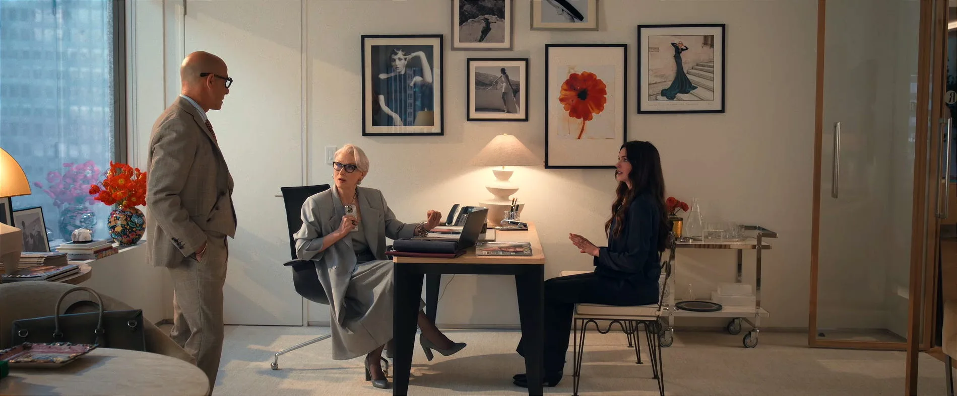 A still from The Devil Wears Prada 2, featuring Anne Hathaway as Andy Sachs, Stanley Tucci as Nigel Kipling and Meryl Streep as Miranda Priestly. They are in Miranda's office, which features white walls, framed photographs on the walls and a light timber and black desk with a white lamp on top. A chrome bar cart is to the side.