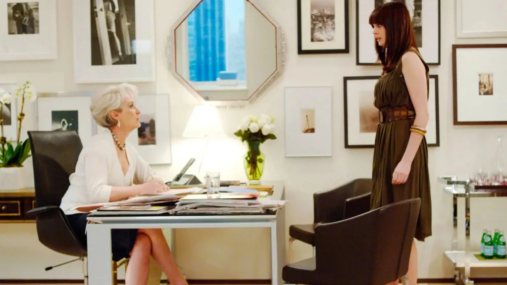A still from the movie The Devil Wears Prada, featuring Anne Hathaway as Andy Sachs and Meryl Streep as Miranda Priestly. They are in Miranda's office, which features white walls, black and white photographs in frames, an octagonal shaped mirror, a black leather chair, chrome drinks trolley and chrome and glass desk. A vase of white flowers is on top of the desk.