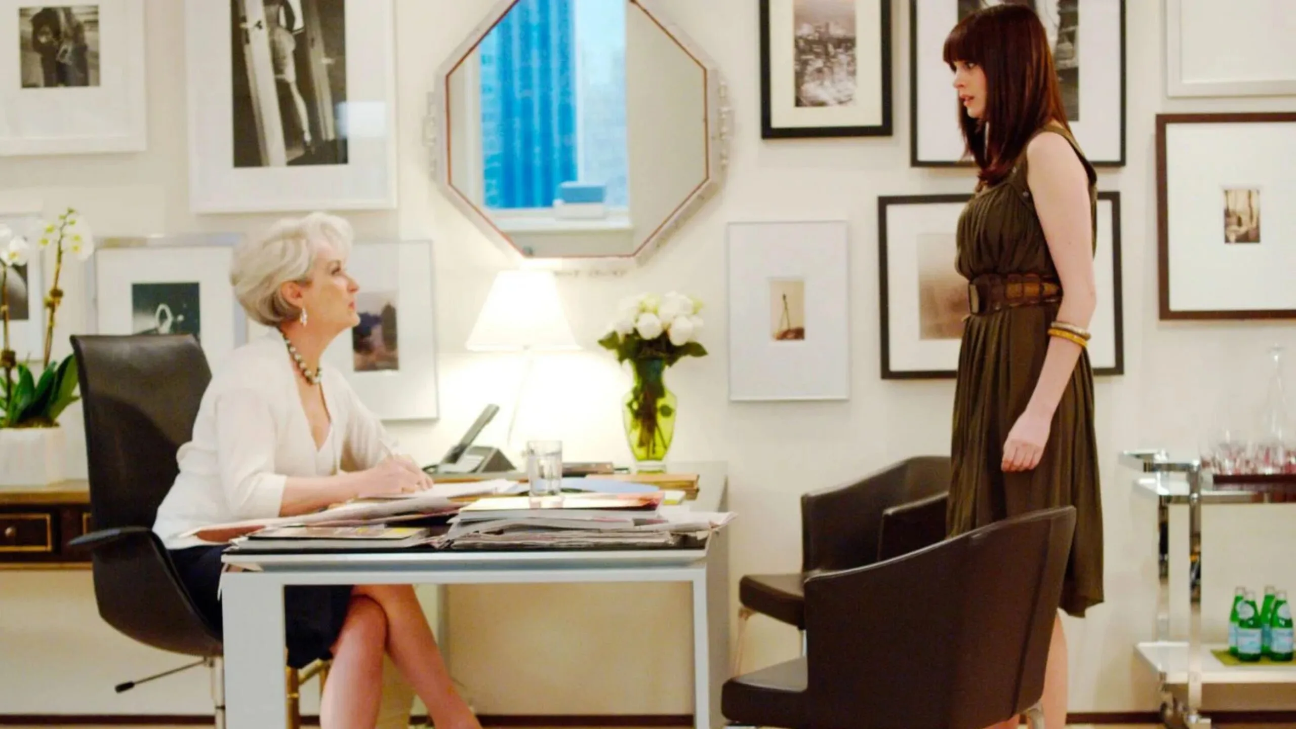 A still from the movie The Devil Wears Prada, featuring Anne Hathaway as Andy Sachs and Meryl Streep as Miranda Priestly. They are in Miranda's office, which features white walls, black and white photographs in frames, an octagonal shaped mirror, a black leather chair, chrome drinks trolley and chrome and glass desk. A vase of white flowers is on top of the desk.