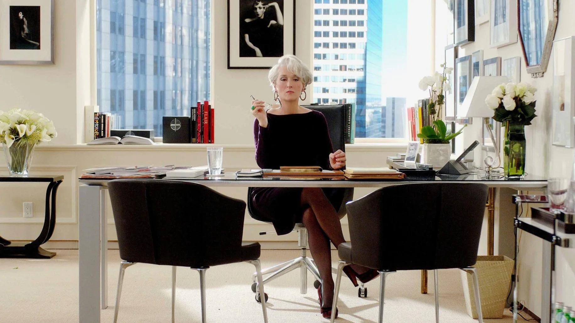 A still from the movie The Devil Wears Prada, featuring Anne Hathaway as Andy Sachs and Meryl Streep as Miranda Priestly. Miranda Priestly is seated behind her glass and chrome desk. The office has white walls decorated with black and white photographs in frames, an octagonal shaped mirror. There's also a black leather chair, chrome drinks trolley and chrome and glass desk. A vase of white flowers is on top of the desk. A view of skyscrapers is visible through the windows behind Miranda.