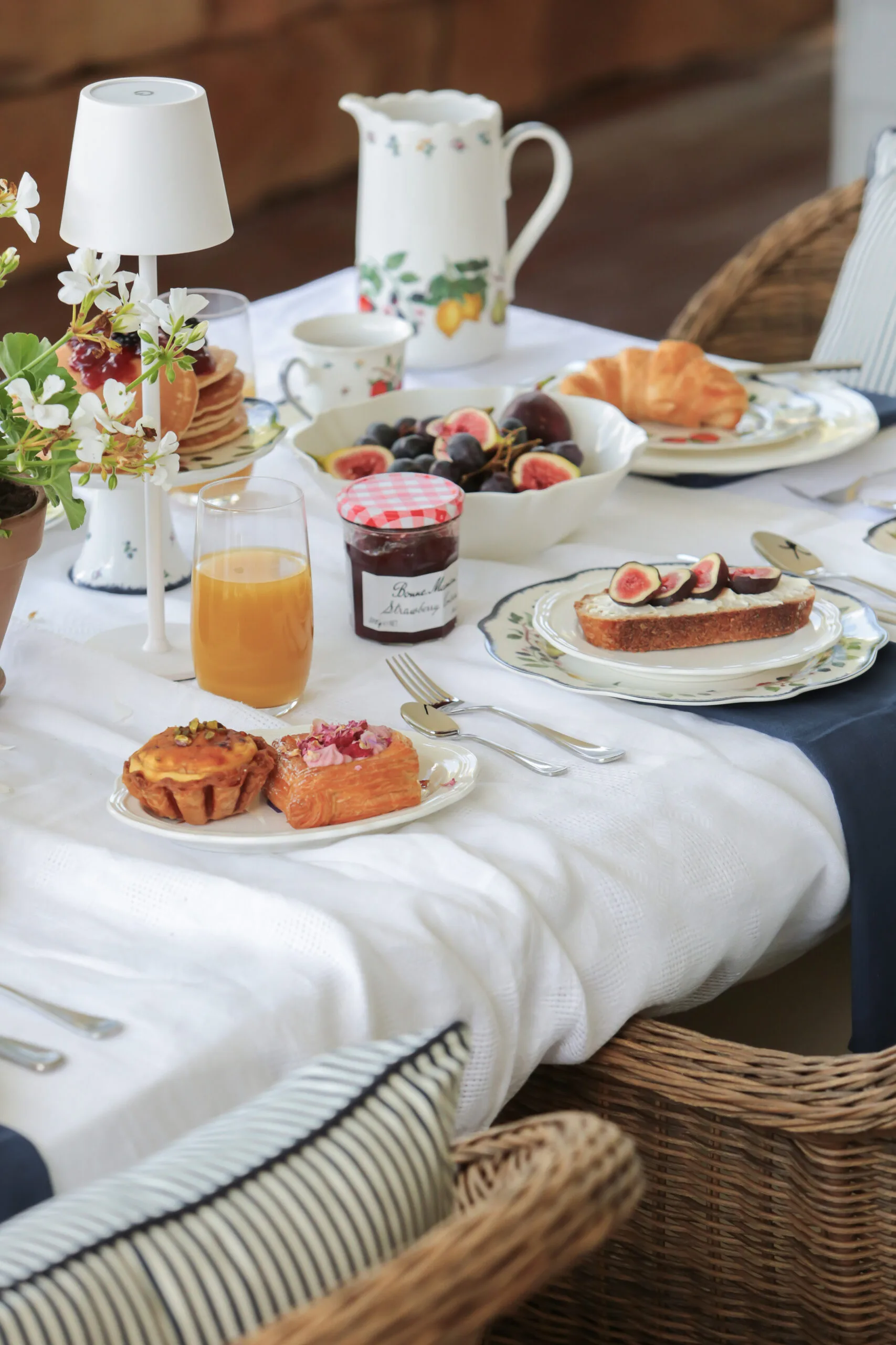 Elegant breakfast table with pastries, figs, orange juice, jam, and floral decor on a white tablecloth.