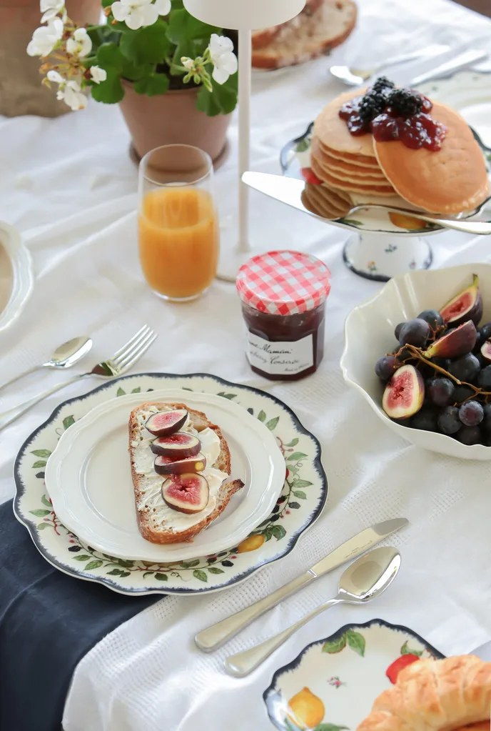 Breakfast table with toast and figs, pancakes with berries, a jar of jam, grapes, orange juice, and flowers.