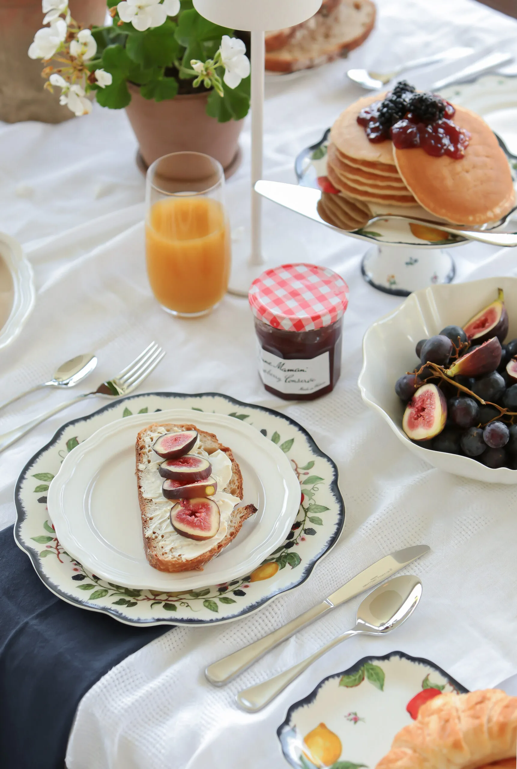 Breakfast table with toast and figs, pancakes with berries, a jar of jam, grapes, orange juice, and flowers.
