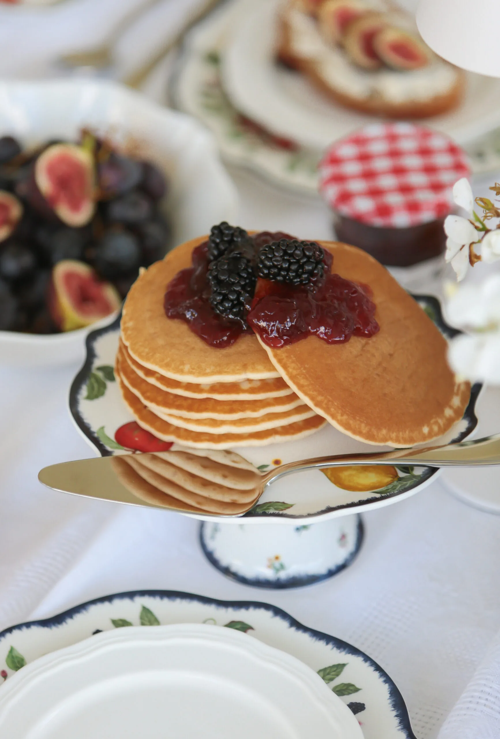 Stack of pancakes with blackberry and jam topping on a floral plate, surrounded by figs and jam jar.