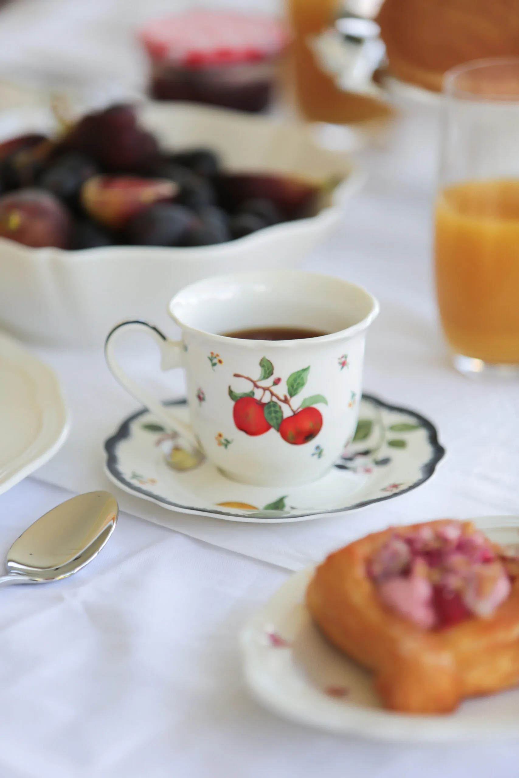 Teacup with cherry design on table, surrounded by fruit bowl, juice glass, and pastry.