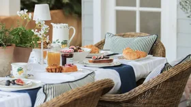Outdoor breakfast setting with pastries, figs, jam, juice, and striped cushions on wicker chairs.