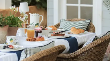 Outdoor breakfast setting with pastries, figs, jam, juice, and striped cushions on wicker chairs.