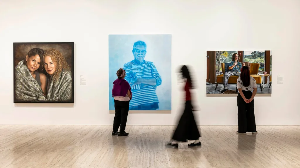 A photo of three people in an art gallery exhibition, one is blurry in motion walking across the floor, the other two are standing in front of portrait paintings.