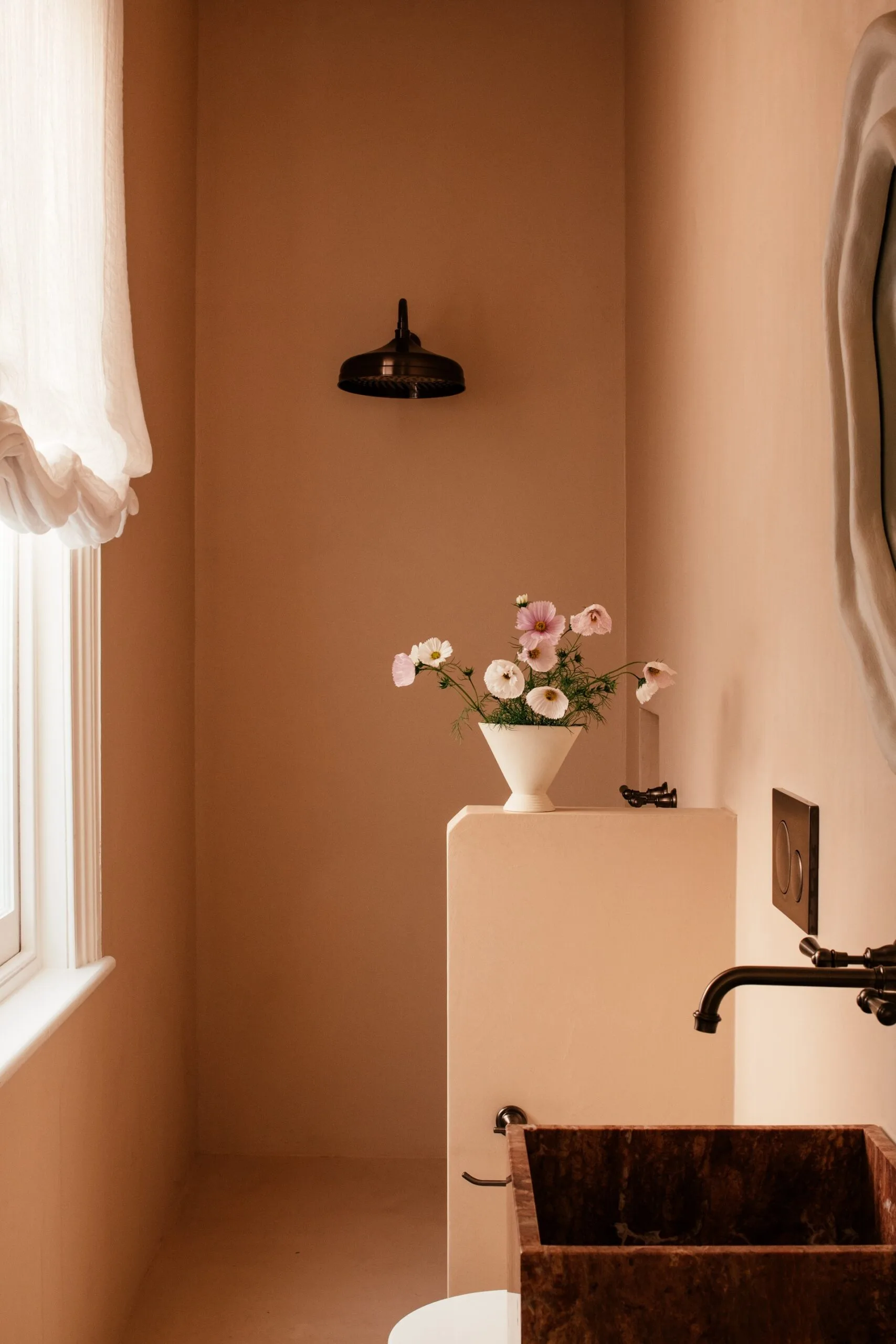 Small ensuite bathroom with pink colour drenched wall and flooring, an aged brass showerhead, marble sink, and vase of pink flowers positioned atop a nib wall, near a window with sheer Roman blinds.