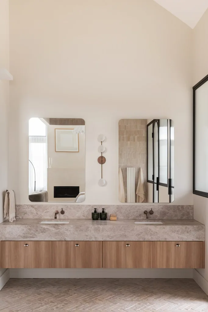 Dual-sink bathroom vanity with marble top, wooden cabinets, and large mirrors above, in a bright, modern setting.