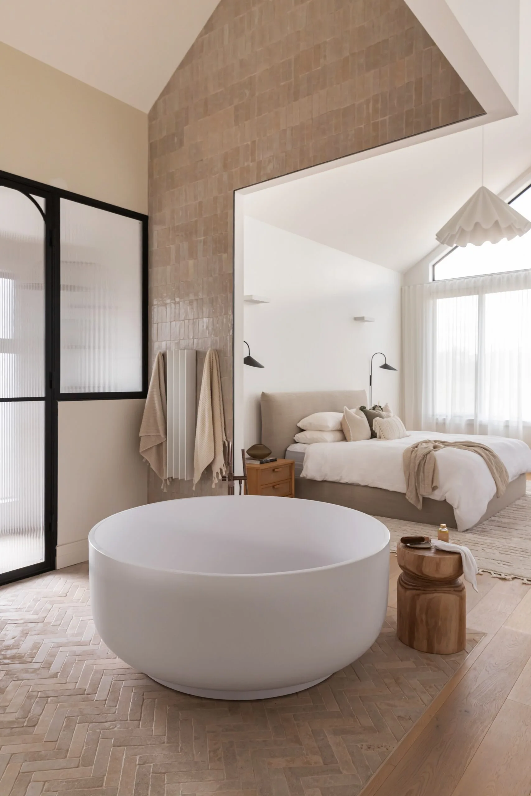 Modern open plan bedroom and ensuite bathroom with neutral tones, featuring a white round bathtub and cozy bedding, accented by natural light.