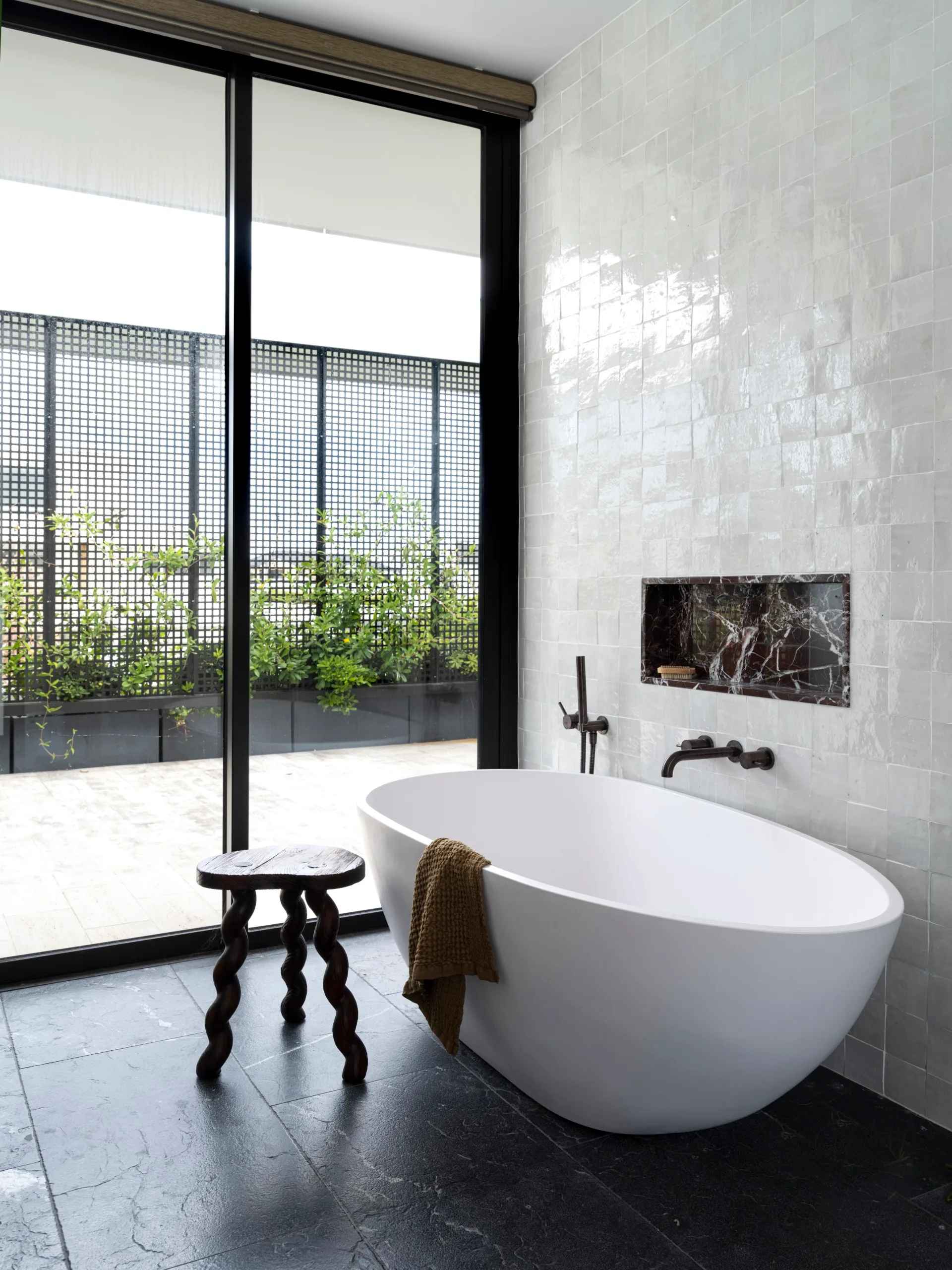 Luxurious bathroom with freestanding white tub, towel draped over edge, modern fixtures, and tall glass doors. A stool is beside the bathtub and a marble niche is built into the wall above the bathtub. A large floor to ceiling door leads on to a balcony, screened from view with plants.