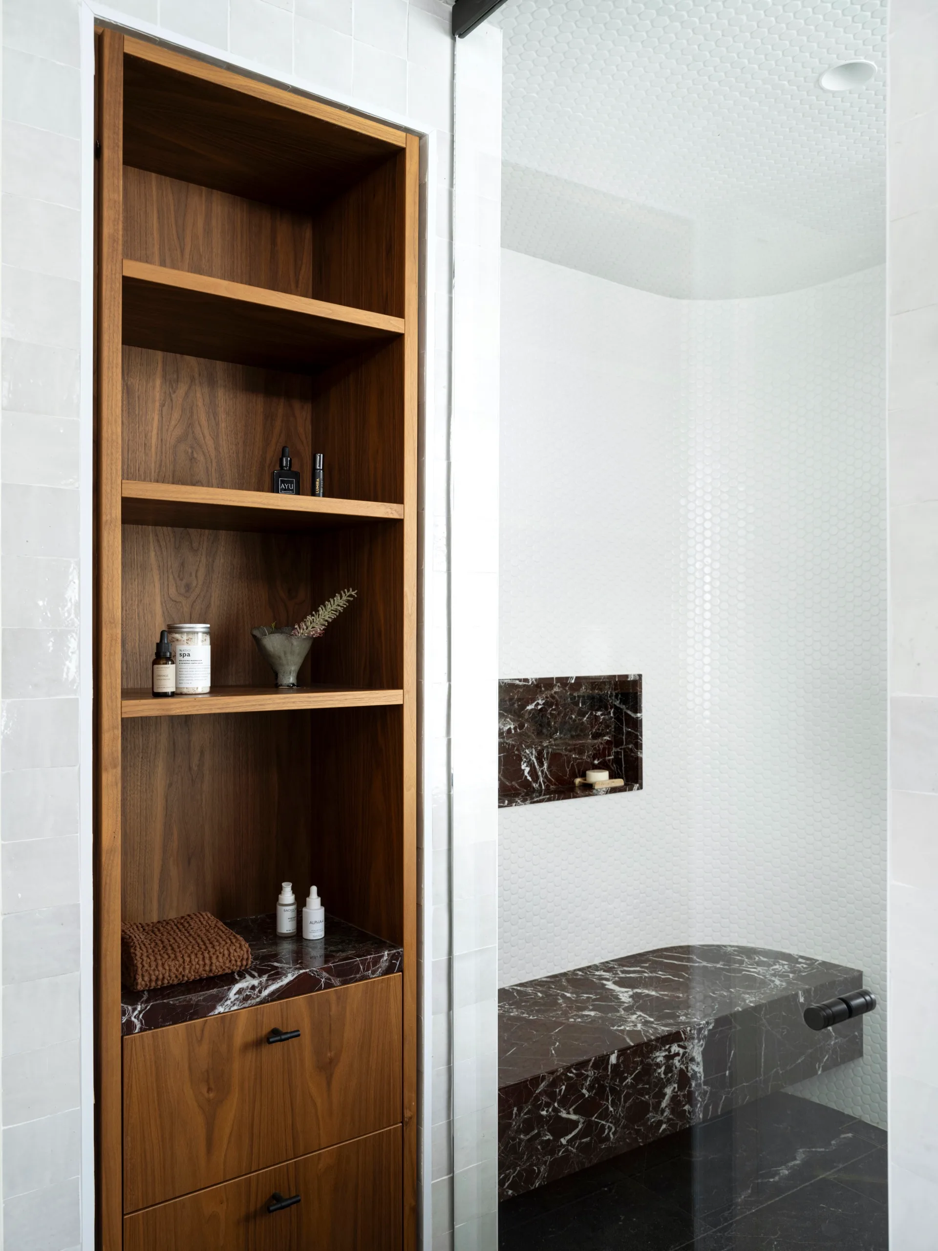 Ensuite bathroom with shower area behind a glass door. A marble shower bench and niche is in the shower area, with penny round white tiled walls. Outside the shower area is wooden open shelving.