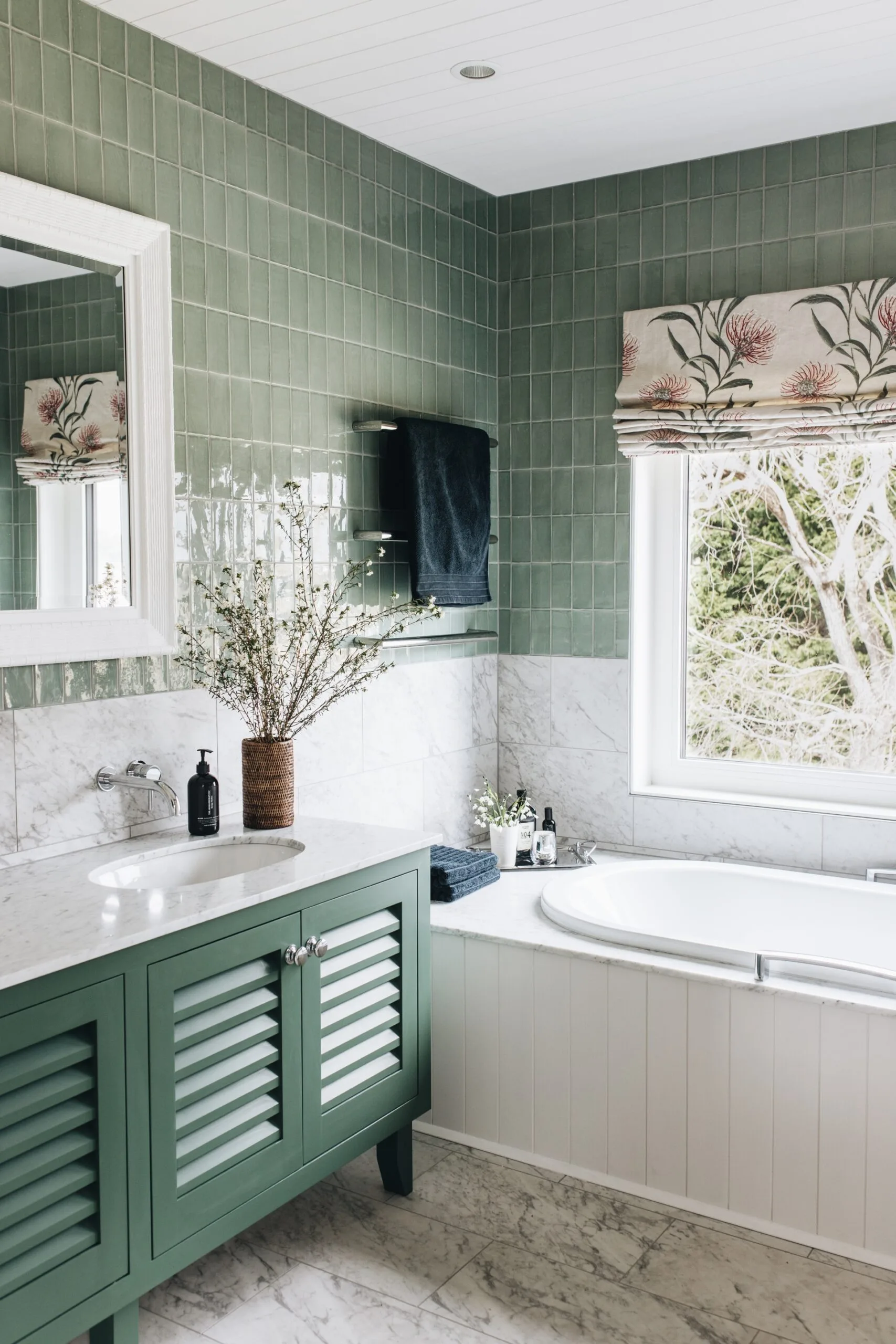 Elegant bathroom with green wall tiles, marble vanity countertop, and a built-in bathtub beside a window with floral blinds. The vanity joinery is also green.