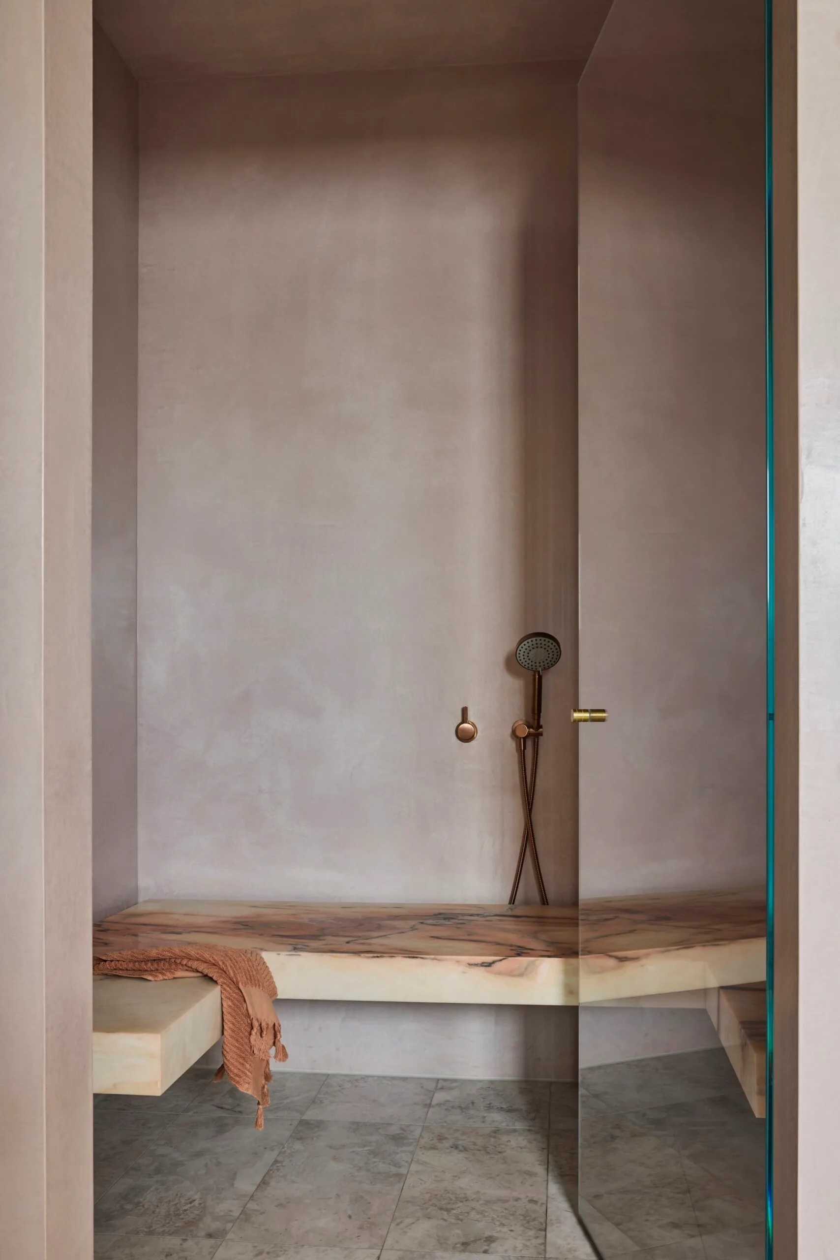 A steam room with glass door, microcement walls finished in a pinkish neutral, and a marble seat.