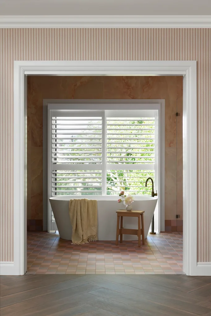 Modern bathroom with freestanding tub, beige towel, wooden stool with flowers, and white shutters overlooking greenery. The tiled floor is a checker pattern of pinks and blues.