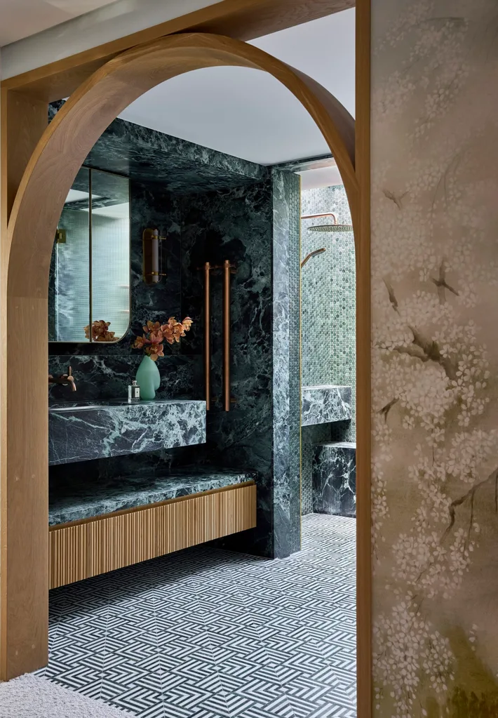 Luxurious bathroom with green marble, wood accents, mosaic tiles, and glass shower doors leading to separate shower and toilet areas. An arch is in the foreground, leading from the bedroom to the bathroom. The room is symmetrical beyond that.