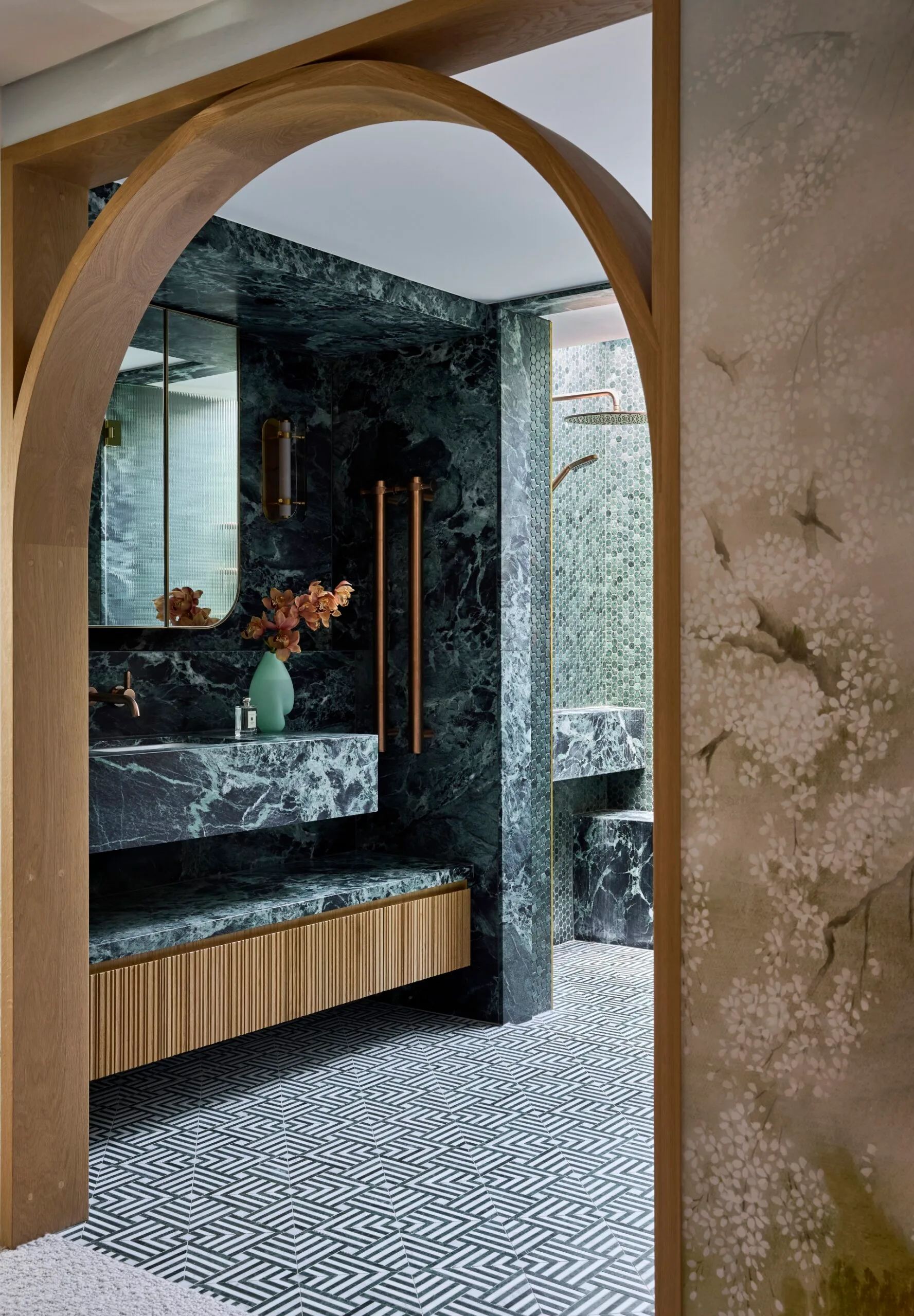 Luxurious bathroom with green marble, wood accents, mosaic tiles, and glass shower doors leading to separate shower and toilet areas. An arch is in the foreground, leading from the bedroom to the bathroom. The room is symmetrical beyond that.