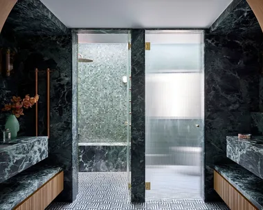 Luxurious bathroom with green marble, wood accents, mosaic tiles, and glass shower doors leading to separate shower and toilet areas. An arch is in the foreground, leading from the bedroom to the bathroom. The room is symmetrical beyond that.